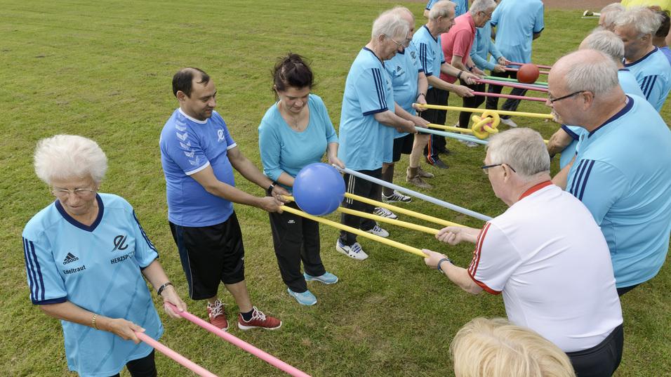 Eine Gruppe &auml;lterer Menschen steht in einer Reihe und h&auml;lt B&auml;lle mit langen bunten St&auml;ben auf einer gr&uuml;nen Wiese.