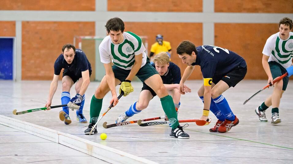 Fünf Spieler in Sportkleidung spielen Hockey in einer Halle, um einen Ball kämpfen sie an einer weißen Linie.