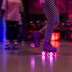 Rollschuhfahrerin mit bunten LED-Rollen in einer sportlichen Umgebung, unscharf hinter einer weiteren Person.