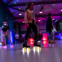 Person in Rollschuhen bewegt sich in einer beleuchteten Halle mit bunten Lichtern und reflektierenden Oberfl&auml;chen.