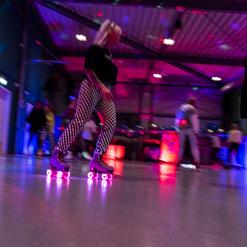 Bewegte Skaterinnen mit bunten Lichtern in einer Sporthalle, die auf Rollschuhen &uuml;ber den gl&auml;nzenden Boden fahren.