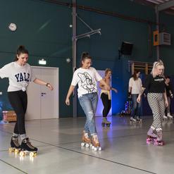 Gruppen von Frauen skaten auf Rollschuhen in einer Halle mit bunter Beleuchtung und DJ-Equipment im Hintergrund.
