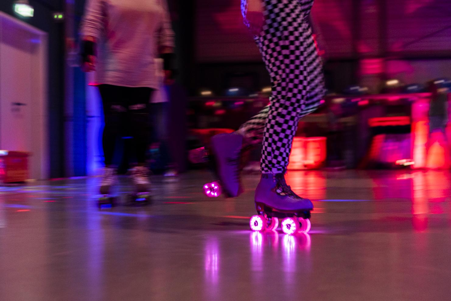 Rollschuhfahrerin mit bunten LED-Rollen in einer sportlichen Umgebung, unscharf hinter einer weiteren Person.
