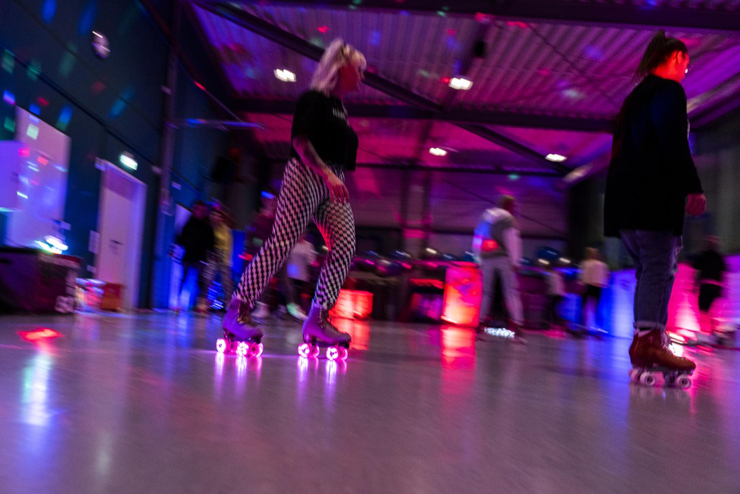 Bewegte Skaterinnen mit bunten Lichtern in einer Sporthalle, die auf Rollschuhen &uuml;ber den gl&auml;nzenden Boden fahren.