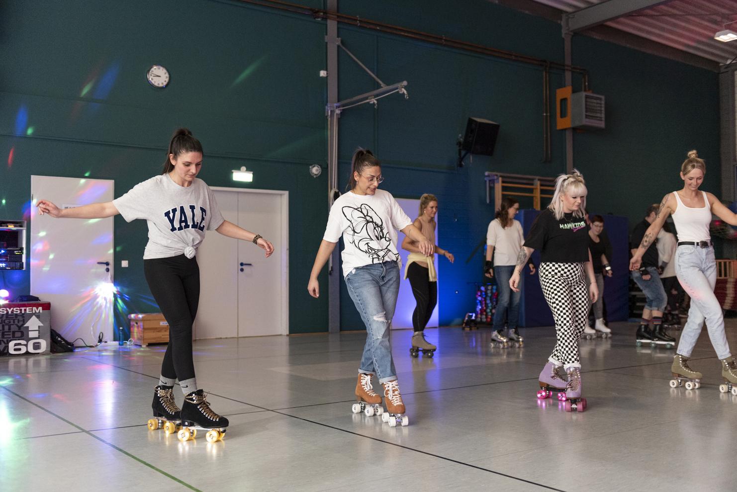 Gruppen von Frauen skaten auf Rollschuhen in einer Halle mit bunter Beleuchtung und DJ-Equipment im Hintergrund.