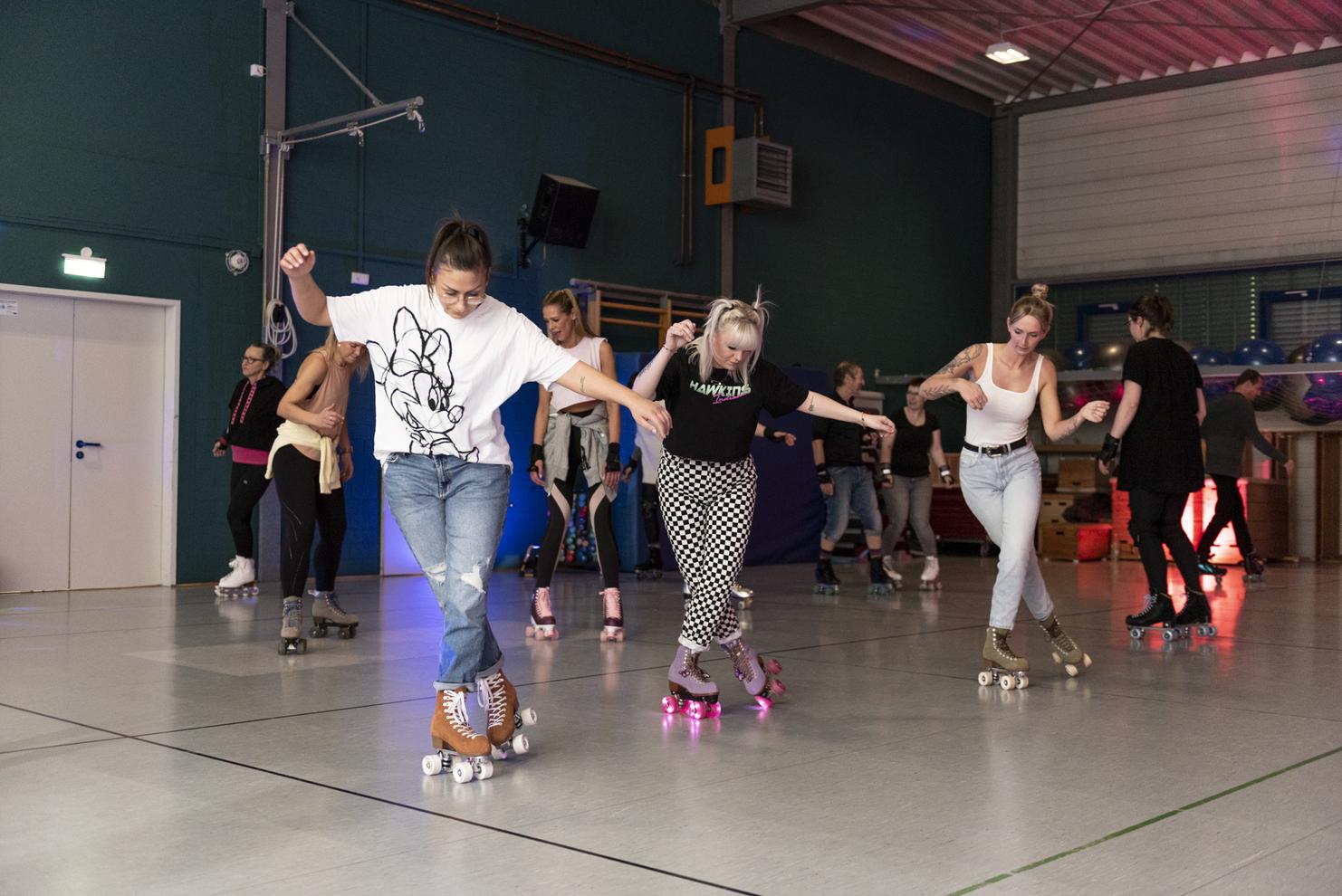 Gruppe von Menschen auf Rollschuhen in einer Sporthalle, die synchron auf einer Fl&auml;che gleiten.