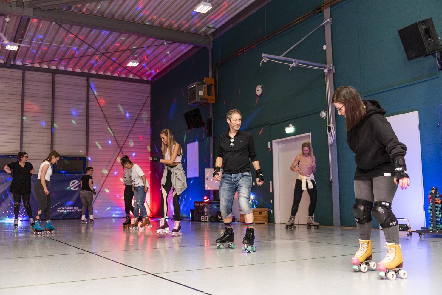Gruppen von Menschen skaten auf Rollschuhen in einer beleuchteten Halle mit bunten Lichtern und reflektierendem Boden.