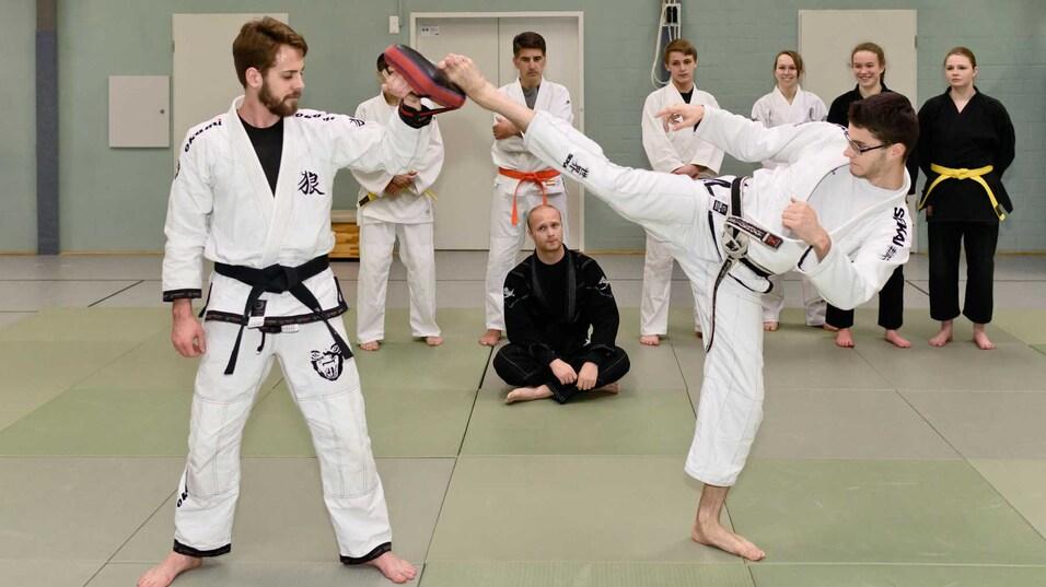 Zwei Kampfsportler in Trainingsanz&uuml;gen &uuml;ben einen Kick an einem Pratze, w&auml;hrend andere Teilnehmer zuschauen.