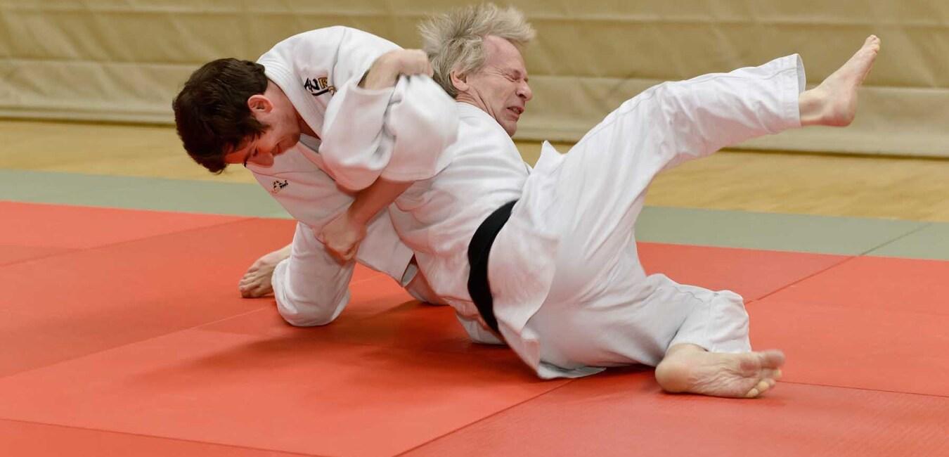 Zwei Judoka in Wettkampfposition auf einer roten Judomatte, einer führt einen Wurf aus.