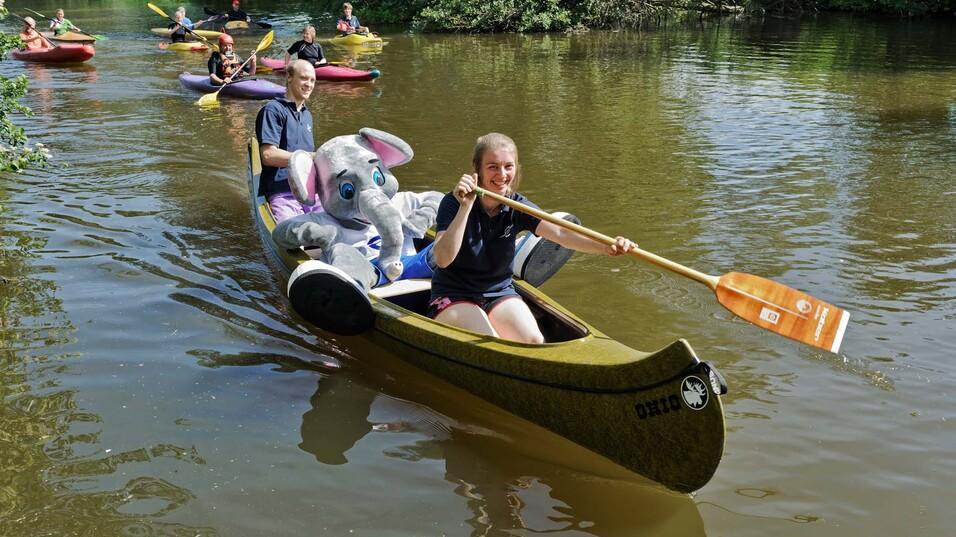 Gruppe von Kanufahrern auf einem ruhigen Gewässer, ein Mädchen paddelt mit einem Plüschelefanten im Boot.