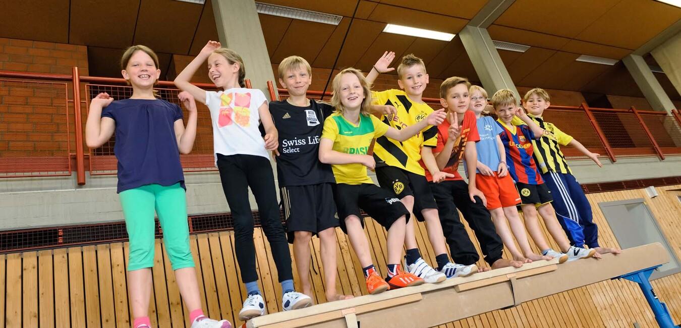 Gruppe von Kindern in Sporttrikots steht auf einer Kline und posiert fr&ouml;hlich in einer Sporthalle.