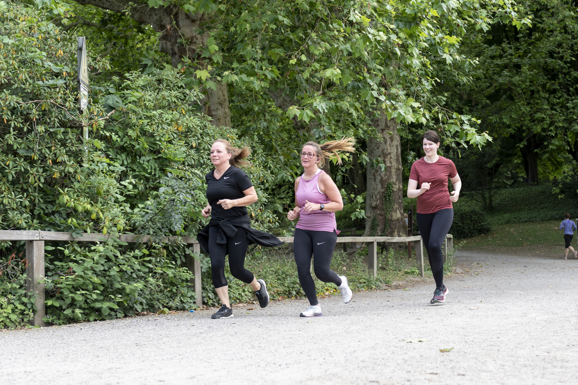 Drei Frauen joggen auf einem Kiesweg, umgeben von gr&uuml;nen B&auml;umen und Str&auml;uchern im Park.