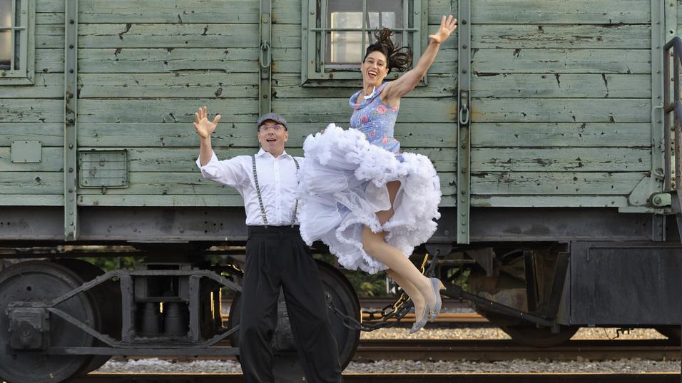 Tanzendes Paar in Vintage-Outfits springt vor einem alten G&uuml;terwagen mit einem fr&ouml;hlichen Ausdruck.