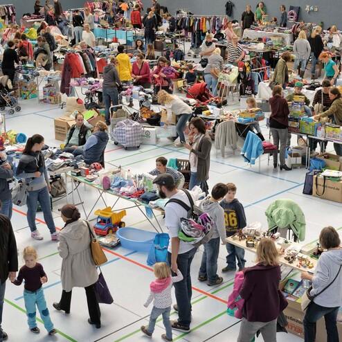 Menschen st&ouml;bern in einem lebhaften Flohmarkt mit vielen St&auml;nden voller Kleidung und Gegenst&auml;nde in einer Halle.