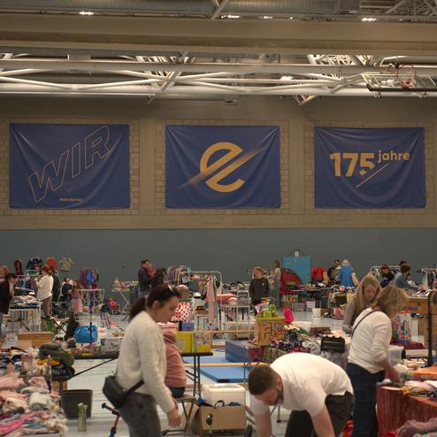 Blick auf einen Flohmarkt in einer Halle mit St&auml;nden voller Kleidung und Interessierten, darunter zwei Frauen beim St&ouml;bern.