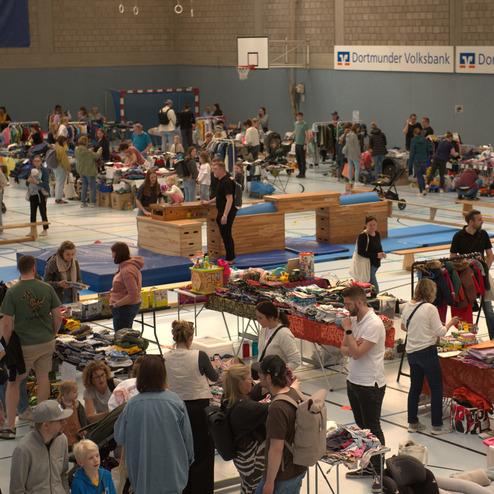 Blick in eine belebte Sporthalle mit St&auml;nden, Menschen und verschiedenen Second-Hand-Artikeln.