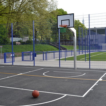 Basketballkorb auf einem Spielfeld mit einem Basketball vor dem Korb und einem geschlossenen Zaun im Hintergrund.