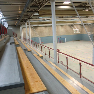 Leere Zuschauertrib&uuml;ne in einer Sporthalle mit Blick auf das Halleninnere und Basketballkorb im Hintergrund.