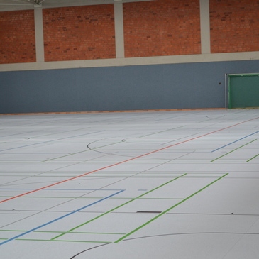 Gro&szlig;e Sporthalle mit hellgrauem Boden, farbigen Linien f&uuml;r verschiedene Sportarten und gr&uuml;nem Tor am Wandende.