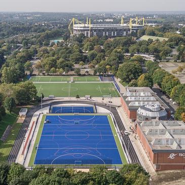 Luftaufnahme eines Sportgeländes mit einem blauen Kunstrasenplatz und einem Fußballfeld im Hintergrund. Stadion sichtbar.