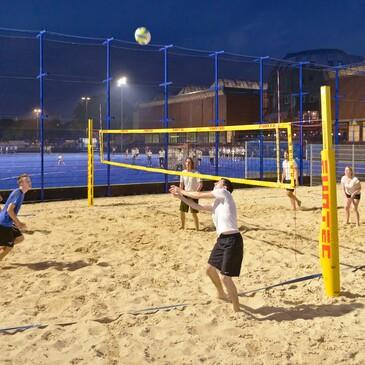 Sechs Personen spielen Beachvolleyball auf einem Sandplatz unter künstlicher Beleuchtung in der Abenddämmerung.