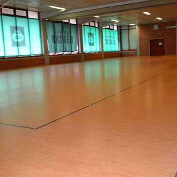 Leere Sporthalle mit Holzfu&szlig;boden und gro&szlig;en Fenstern, die von gr&uuml;nen Vorh&auml;ngen umrahmt sind.