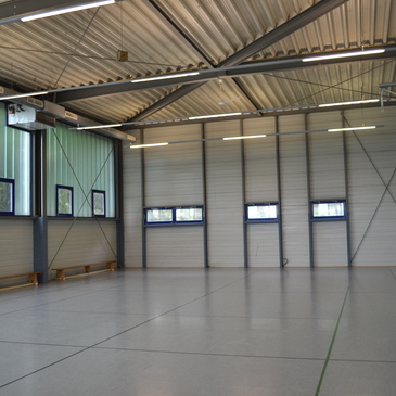Leere Sporthalle mit grauem Boden, hohen Decken und gro&szlig;en Fenstern auf der linken Seite.