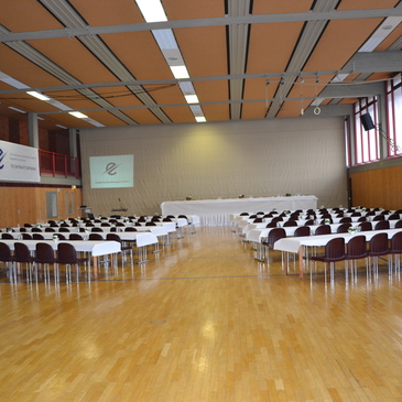Blick in einen gro&szlig;en Saal mit Reihen von Tischen und St&uuml;hlen f&uuml;r eine Veranstaltung, vorne eine Leinwand.