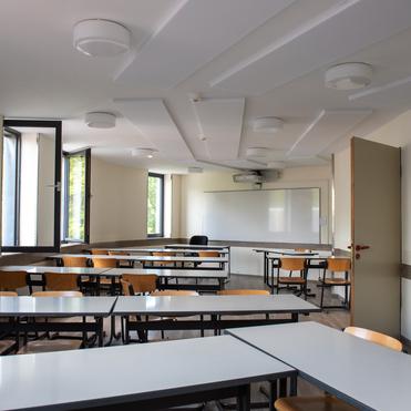 Helle Klassenzimmeransicht mit Tischen, Stühlen und einer Präsentationswand. Fenster sorgen für natürliche Beleuchtung.