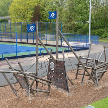 Spielplatz mit Klettergerüst aus Holz und angrenzendem Sportplatz, umgeben von Bäumen und Rasenflächen.