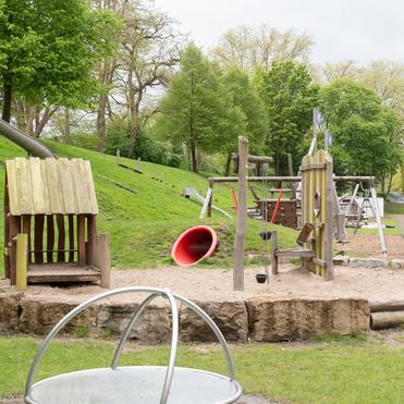 Spielplatz mit Holzspielgeräten, Rutsche und Sandbereich, umgeben von Wiese und Bäumen unter bewölktem Himmel.