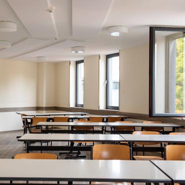 Leerer Klassenraum mit Holzstühlen und Tischen, großen Fenstern und hellen Wänden.