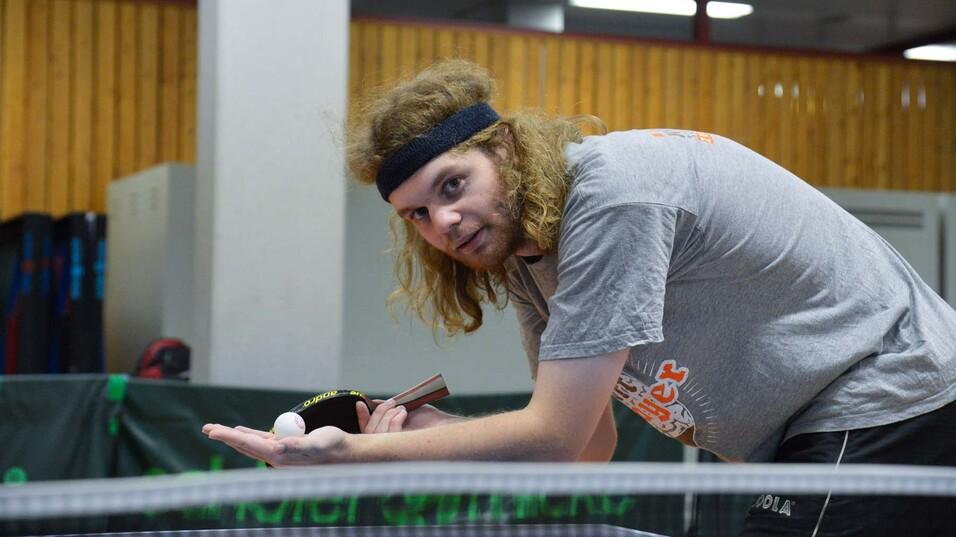 Junger Mann mit lockigen, langen Haaren und Stirnband bereitet sich am Tischtennistisch mit Ball und Schläger auf den Aufschlag vor.
