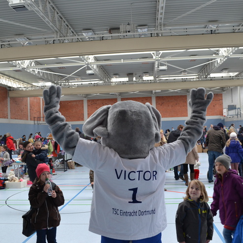 Maskottchen Victor, ein Elefant, steht in einer &uuml;berf&uuml;llten Halle und hebt beide Arme zur Begr&uuml;&szlig;ung der Zuschauer.