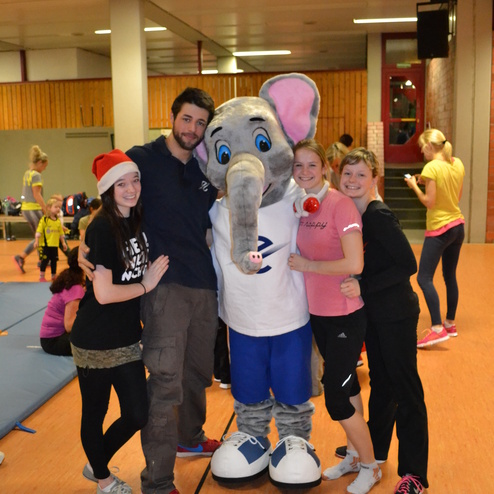 Vier Personen posieren in einem Sportsaal mit einem Elefanten-Maskottchen in Sportkleidung.
