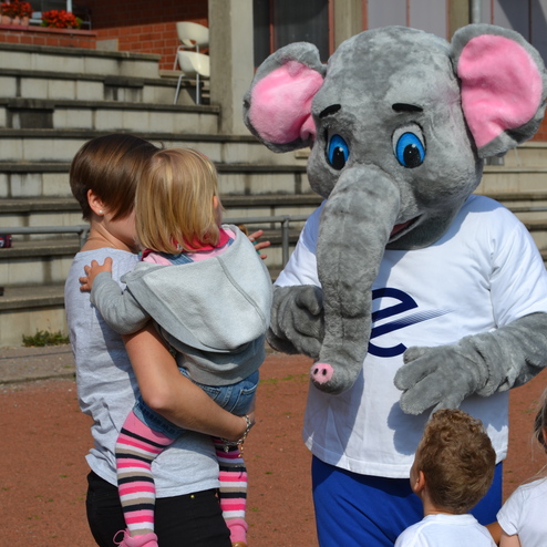 Elefantenmaskottchen interagiert mit Kindern auf einem Sportplatz, w&auml;hrend Erwachsene im Hintergrund sitzen.