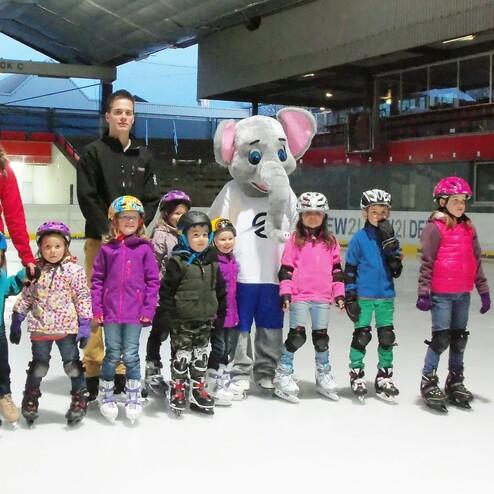 Gruppe von Kindern in Skaterkleidung und zwei Erwachsenen mit einem Maskottchen auf einer Eisfl&auml;che.