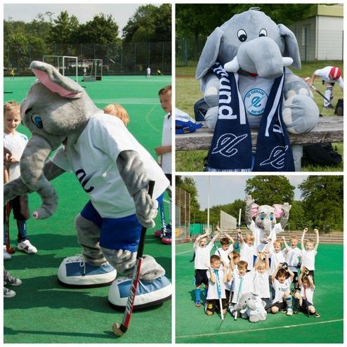 Maske eines Elefanten in Sportkleidung auf einem Hockeyfeld, umgeben von Kindern in Wei&szlig;, die jubeln.