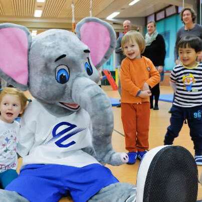 Kindergartenkinder stehen um ein Elefanten-Maskottchen, das auf dem Boden sitzt, w&auml;hrend sie spielen und lachen.