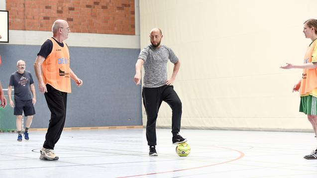 Fünf Männer spielen in einer Sporthalle Fußball, einer kontrolliert den Ball, andere stehen nah daneben.