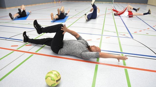 Gruppentraining in einer Sporthalle: Personen liegen auf dem Boden und machen Übungen, ein Fußball liegt daneben.