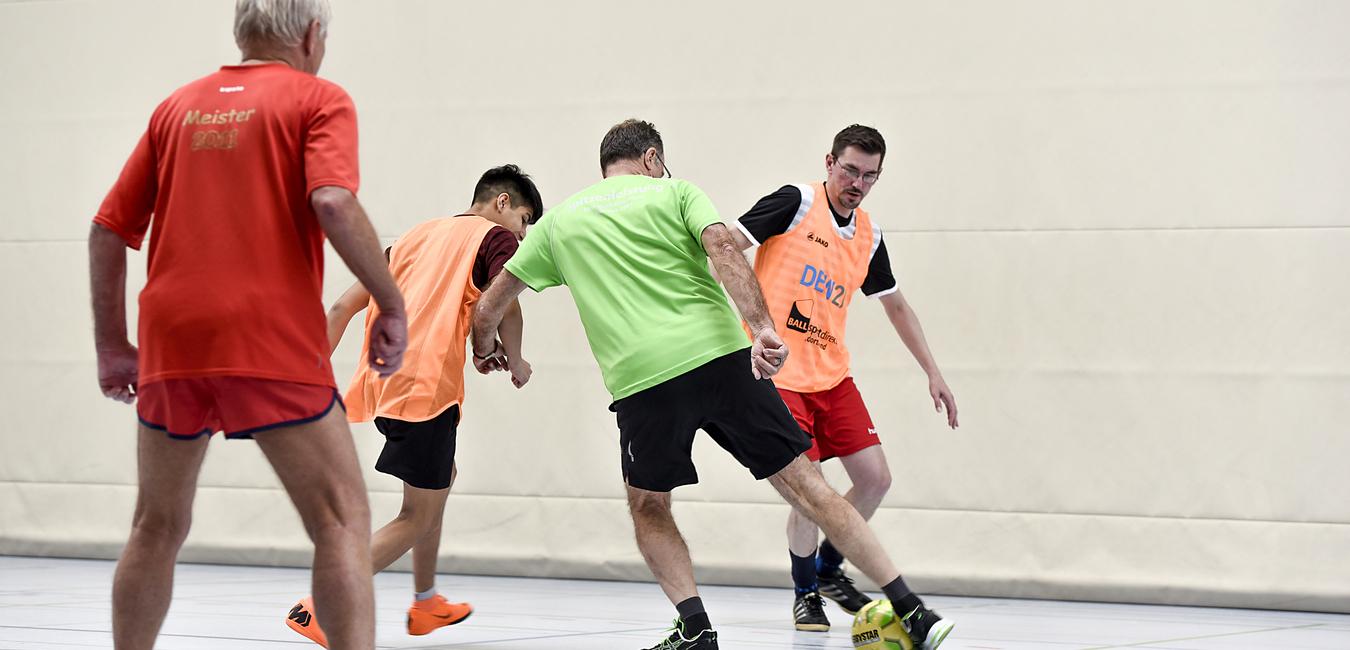 Vier Männer in Sportbekleidung spielen Fußball in einer Halle, ein Spieler trägt ein grünes Trikot und ist am Dribbeln.