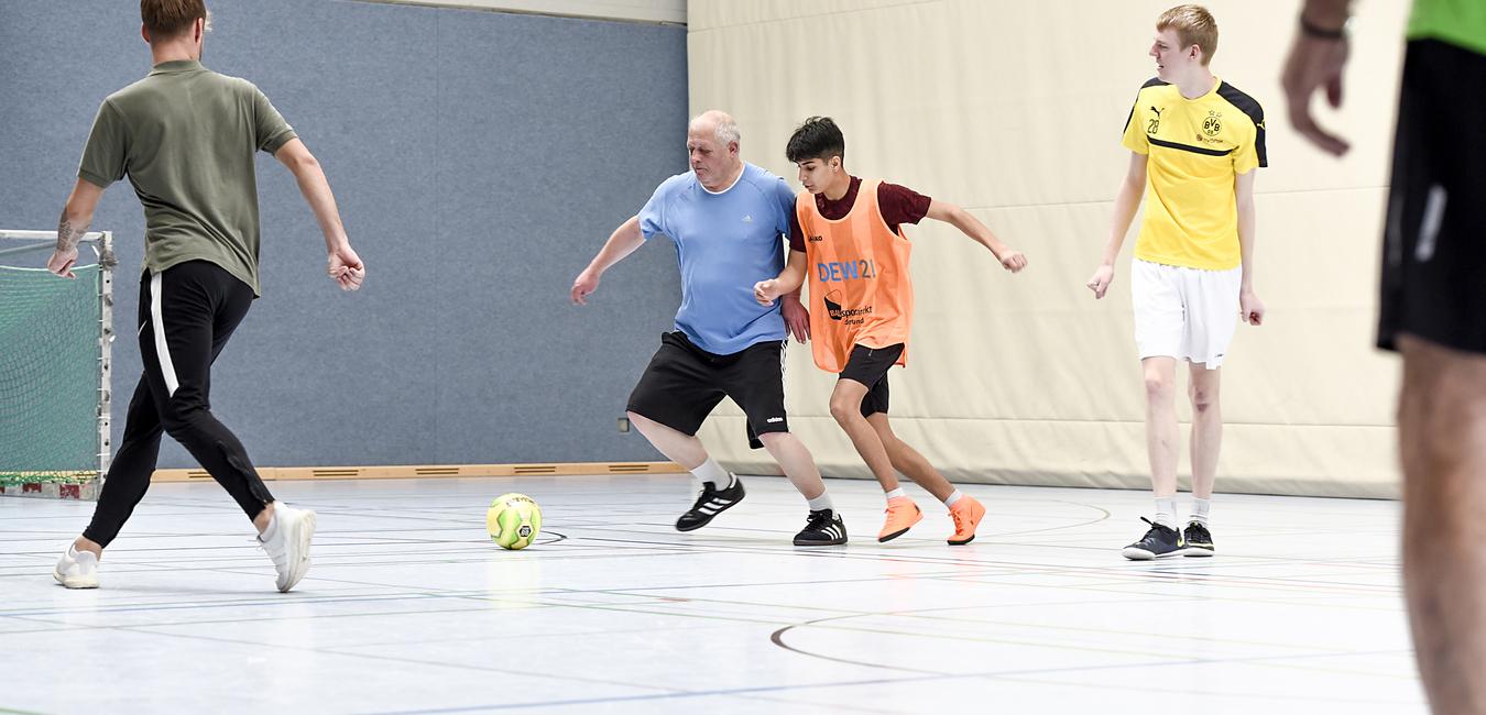 Szenen einer Sporthalle mit mehreren Personen, die intensiv Fußball spielen, während ein Ball in der Mitte liegt.