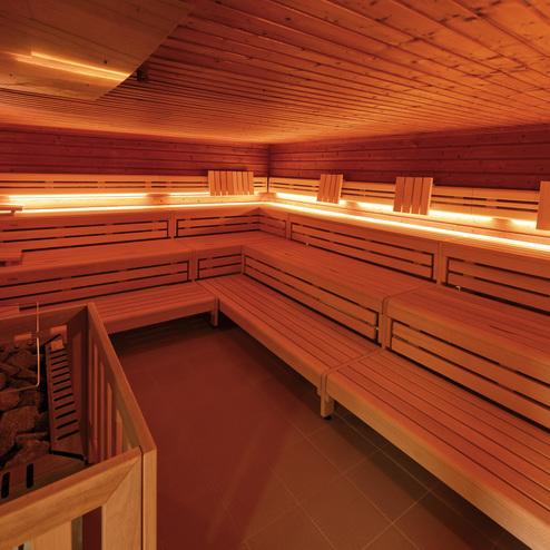 Innenansicht einer Holzsauna mit beleuchteten Bänken und Saunasteinofen auf der linken Seite.