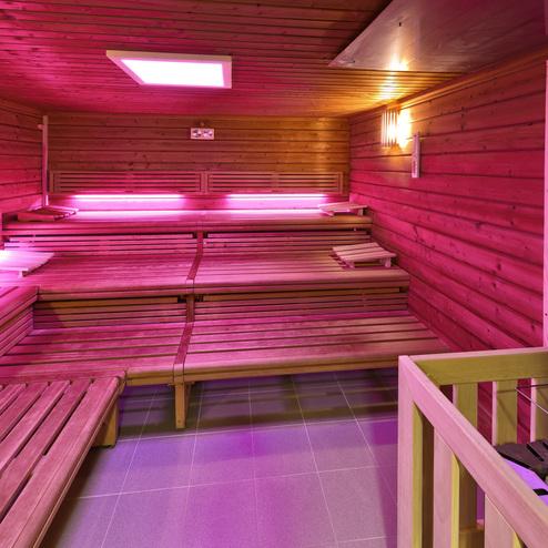 Innenansicht einer Sauna mit Holzbänken und lila Beleuchtung, minimalistischem Design und Steinen am Heizgerät.