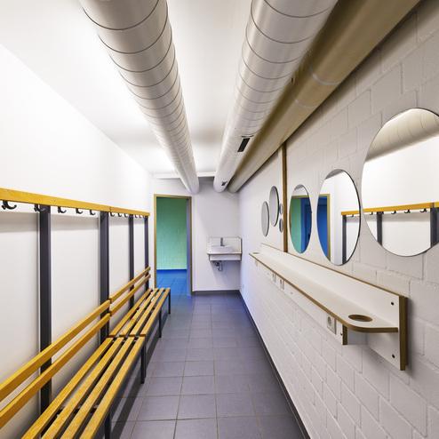 Schlauchförmiger Umkleidebereich mit Holzbänken, Haken und runden Spiegeln an der weißen Wand.