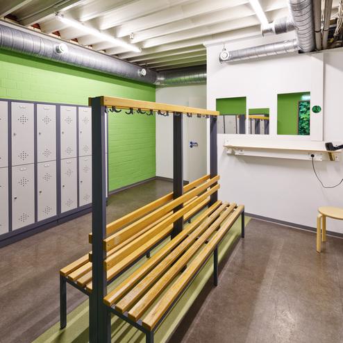 Umkleideraum mit einer Holzbank, Spinden und einem Tisch mit Steckdose an einer grünen Wand.