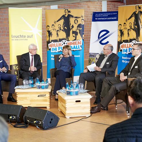 Podiumsdiskussion mit f&uuml;nf Personen auf einem Event, umgeben von Bannern, mit Wassergl&auml;sern auf einer Holzplanke.