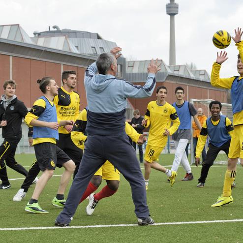 Gruppe von Fu&szlig;ballspielern in gelben Trikots, die auf einem Platz ein Spiel &uuml;ben, w&auml;hrend ein Trainer Anweisungen gibt.