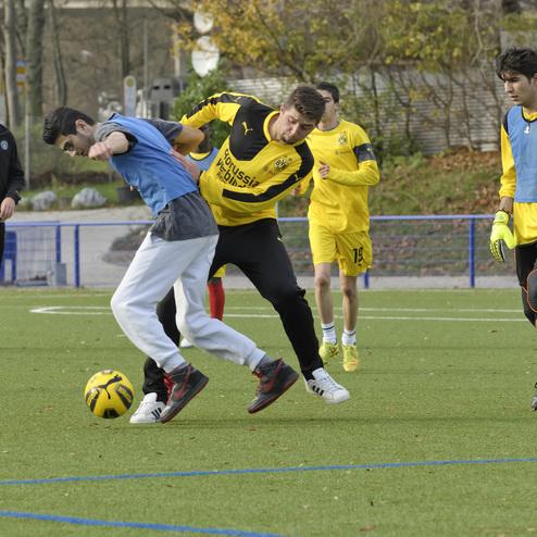 F&uuml;nf Jugendliche spielen Fu&szlig;ball auf einem Kunstrasenplatz, w&auml;hrend ein Spieler den Ball dribbelt.